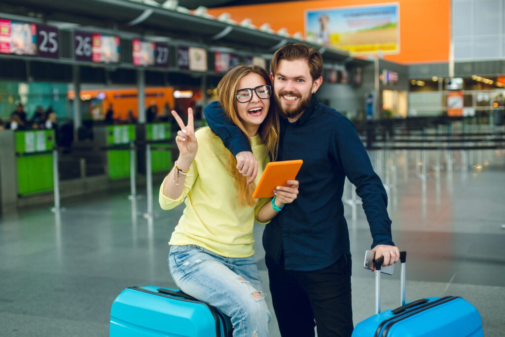 Portrait of young couple hugging in airport. She has long hair, yellow sweater, jeans and tablet. He has black shirt, pants and suitcase near. They are smiling to the camera.