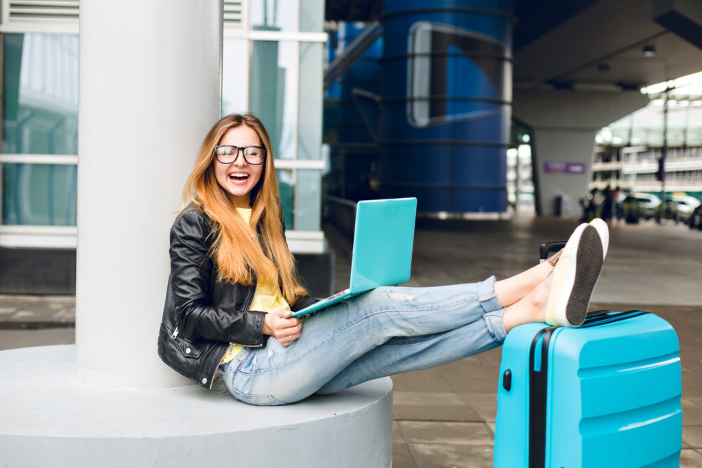 Pretty girl with long hair in glasses is sitting outside in airport. She wears jeans, black jacket, jellow shoes and has laptop. She put her legs on suitcase and smiling to the camera.