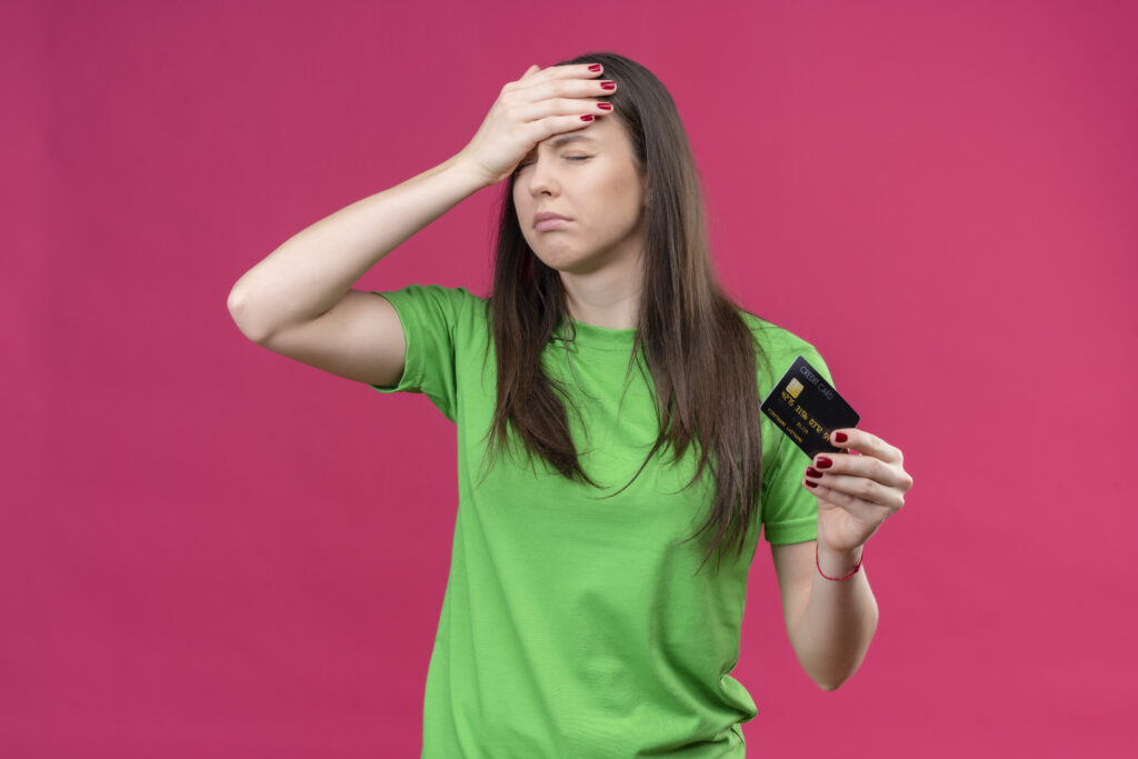 young beautiful girl wearing green t-shirt holding credit card holding her head with hand for mistake upset with closed eyes standing over isolated pink background